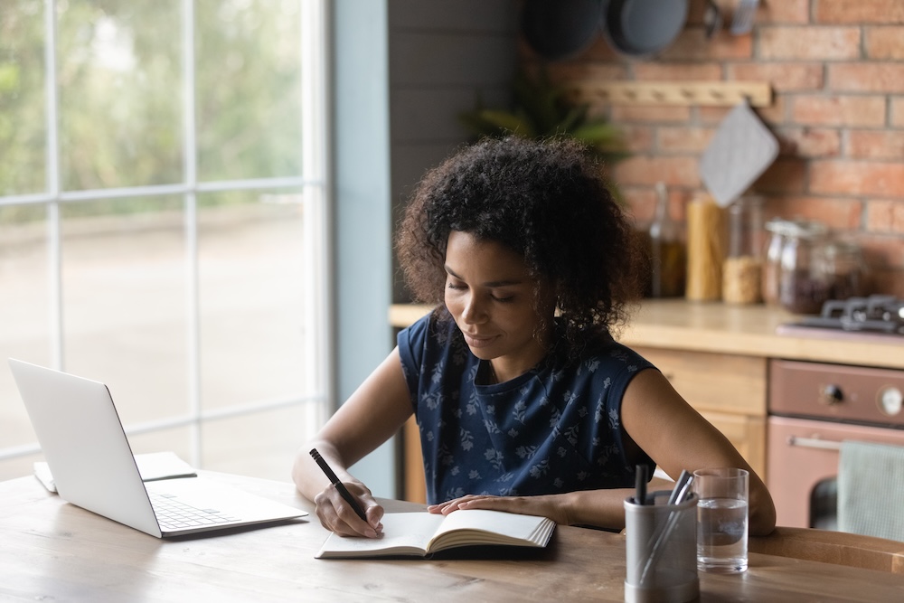 A woman with curly hair sits at a wooden table in a cozy kitchen, jotting notes for WordPress website maintenance. A laptop is open nearby, ready for updates, and a glass of water rests on the table. Large windows bathe the room in natural light, with kitchenware visible in the background.