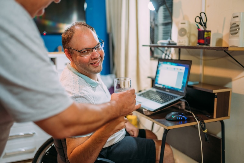 A man in a wheelchair is sitting at a desk with an open laptop, smiling while another person is handing him a glass of water.