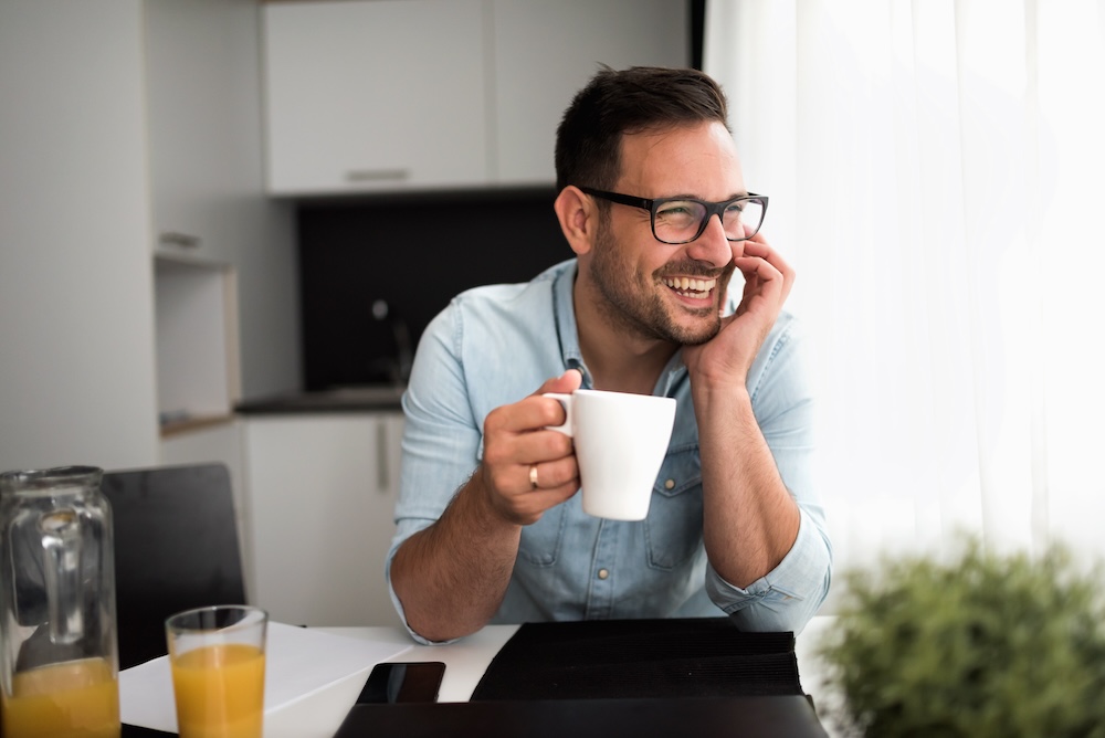 A smiling man with glasses sits at a table, holding a white mug, blissfully overseeing his WordPress website maintenance. He wears a light blue shirt amidst a bright kitchen. On the table, there's a glass of orange juice and a small plant basking in the natural light filling the room.