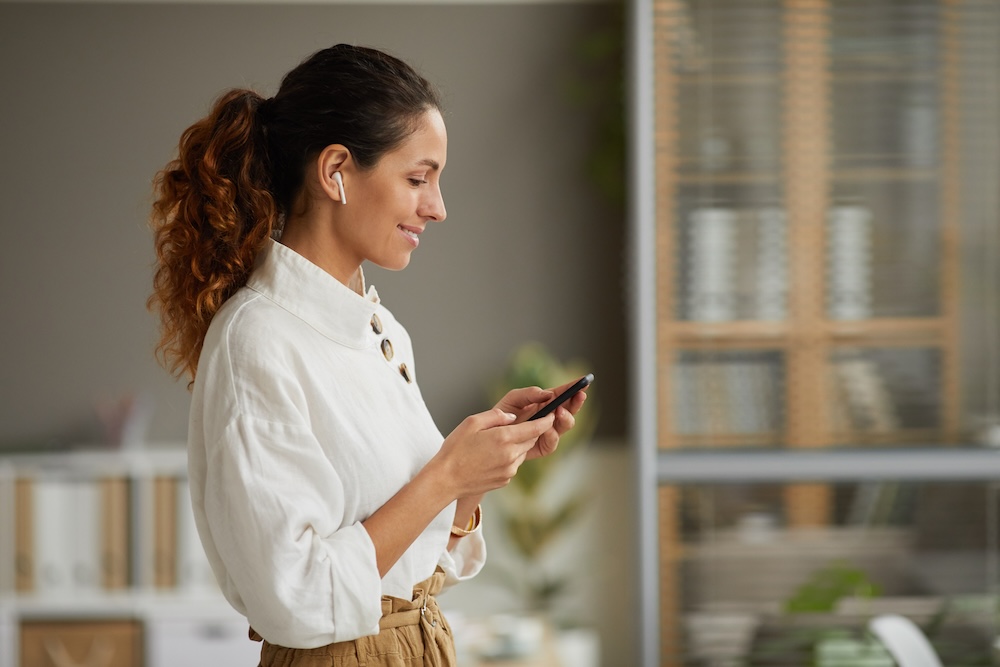 A woman with long hair tied back, wearing a white blouse and wireless earbuds, is smiling while looking at her smartphone. She stands in a modern room with shelves in the background, perhaps reviewing tips on WordPress website maintenance.