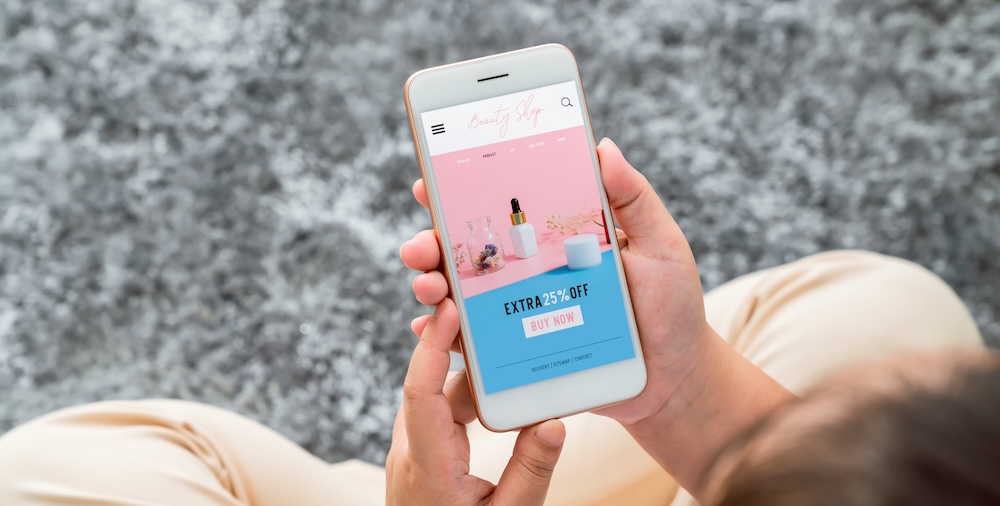 A person relaxes on a gray carpet, smartphone in hand, exploring a beauty product website that's masterfully adapted to a mobile-first world. The vibrant pink background showcases various items with a tempting "25% off" and an inviting "Buy Now" button.