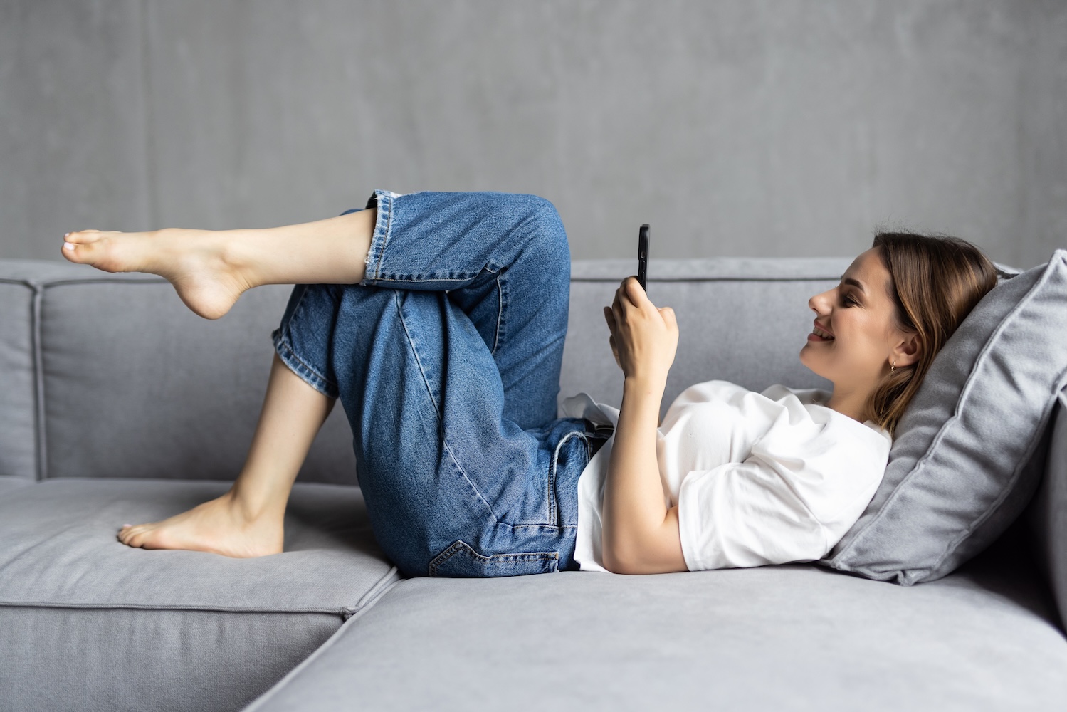 A woman in jeans and a white t-shirt lies on a gray couch, relaxed and smiling while browsing about adapting web design to a mobile-first world on her smartphone. Her legs are propped up on the armrest, enjoying her casual downtime against the plain gray wall.